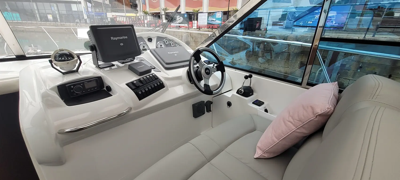 Modern boat cockpit featuring a steering wheel, Raymarine display, controls, and a comfortable seat with a pink cushion.